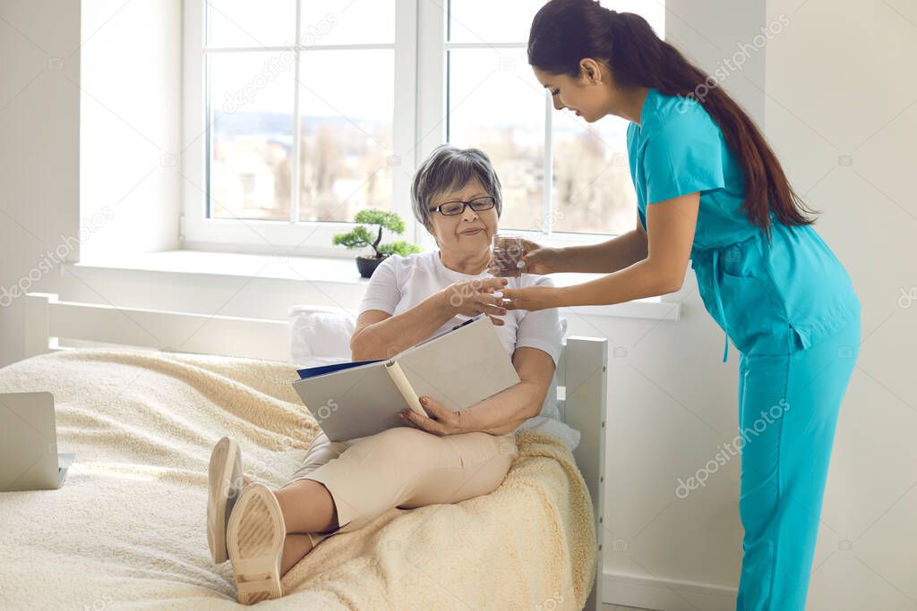 Woman nurse giving prescribed pills and glass of water to happy elderly lady