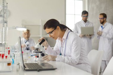 Konsantre kadın virolog laboratuarda masada oturur ve mikroskop altında hücreleri inceler..