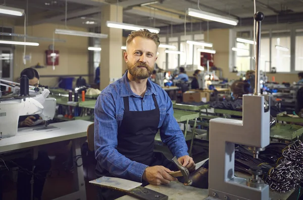 Retrato de um homem amigável sentado em seu local de trabalho e usando um martelo para fazer cintos em uma fábrica. — Fotografia de Stock