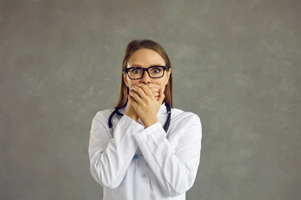 Worried female doctor with a frightened expression covering her mouth on a gray background. — Stock Photo, Image