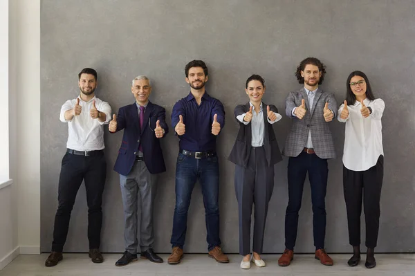 Líderes de negócios mostrando polegares para cima enquanto estão contra a parede cinza e olhando para a câmera. — Fotografia de Stock