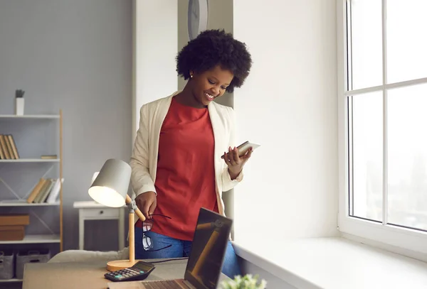 Heureuse jeune femme d'affaires utilisant un téléphone portable debout près de la fenêtre dans son bureau — Photo