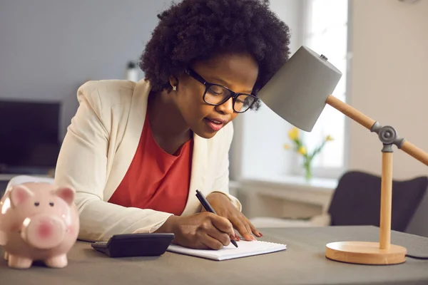 Millénaire afro-américaine femme d'affaires écrit des notes tout en étant assis près de calculatrice et tirelire — Photo