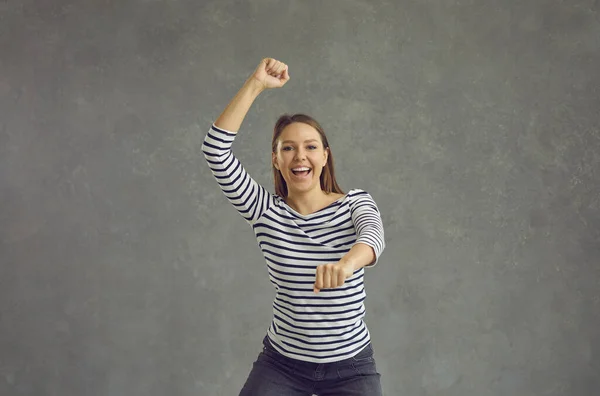 Radostná mladá žena tančí bezstarostně na šedém pozadí slaví vítězství a úspěch. — Stock fotografie