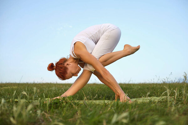 Молодая женщина с гибким телом практикует йогу упражнения на траве против неба.