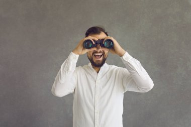 Genç adam gri stüdyo arka planına gülen yüzlü dürbünle bakıyor.