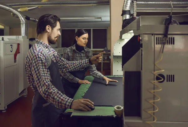 Two employees of a shoe factory work together with a machine for cutting fabric and leather.