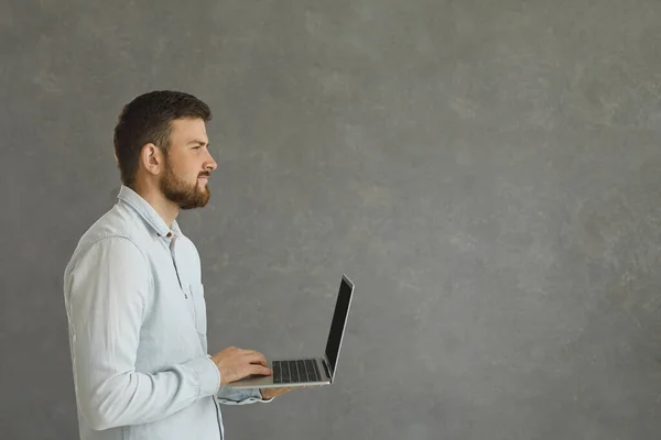 Portrét mladého a sebevědomého muže stojícího na šedém pozadí s laptopem v ruce. — Stock fotografie