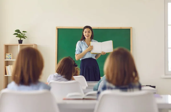 Női tanár áll a táblánál egy könyvvel a kezében fiatal diákok előtt. — Stock Fotó
