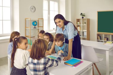 Güler yüzlü beyaz bayan öğretmen çocuklara yardım ediyor ve okul malzemelerini anlatıyor..