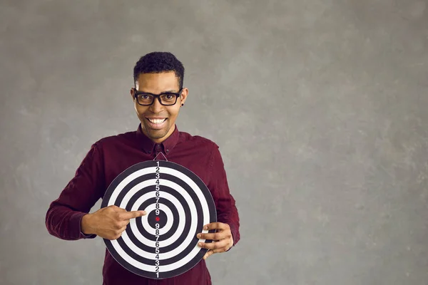 Jonge Afro-Amerikaanse man die een dartbord vasthoudt en naar het midden wijst. — Stockfoto