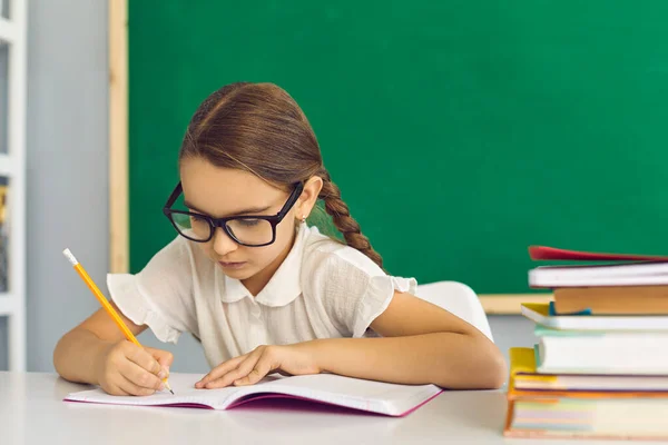 Маленькая студентка сидит за столом в классе и пишет в школьной тетради — стоковое фото