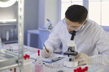 Bilimadamı mikrobiyoloji laboratuvarında mikroskopla masada otururken notlar alıyor.
