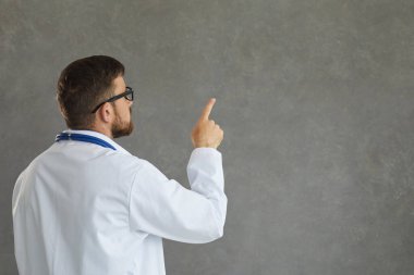 Odaklanmış erkek doktor, sanki gri arka planda bir şey okuyup parmağıyla işaret ediyor gibi..
