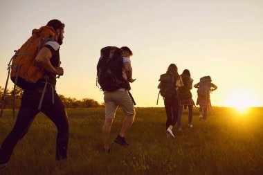 Bir grup turist gün batımında yeşil yaz sahasında sırt çantalarıyla yürüyüş yapıyor.