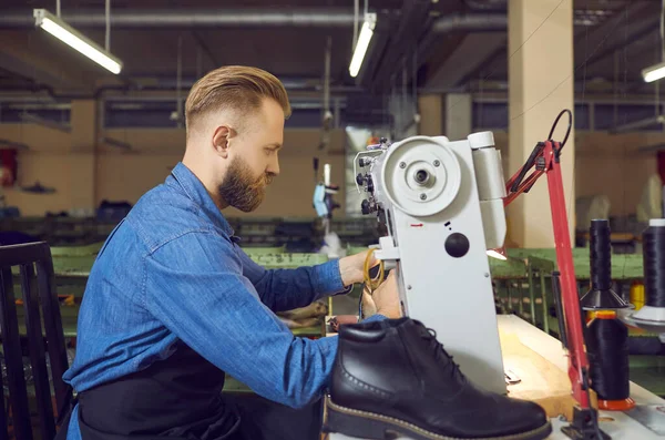 Trabalhador da fábrica de sapatos usando uma máquina de costura industrial para fazer novas botas de couro preto — Fotografia de Stock