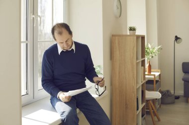 Olgun bir işadamı, ofis penceresinin kenarında oturan kağıtlarla çalışıyor.