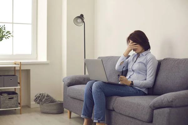 Woman sitting on sofa with laptop stops working and covers face feeling tired and unhappy — Stock Photo, Image