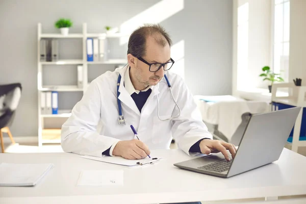 Médico sério usando notas de escrita de laptop em diário médico sentado na mesa — Fotografia de Stock