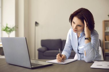 Önünde dizüstü bilgisayarı olan bir kadın oturuyor ve ciddi ciddi deftere yazı yazıyor..