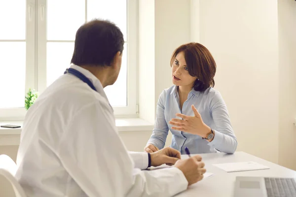 Mulher branca de meia-idade paciente descrevendo sintomas conversando com o médico no consultório — Fotografia de Stock