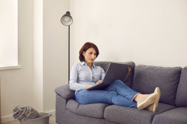 Orta yaşlı beyaz bir kadın kanepede dizüstü bilgisayarda yazarak sohbet ediyor.