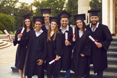 Üniversite kampüsünün yanında diplomaları olan cüppe ve şapkalı yüksek lisans öğrencileri. Gençler mezuniyeti kutluyor