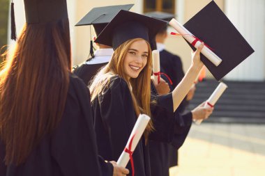 Gülümseyen güzel bir kız üniversite ya da üniversite mezunu diplomasıyla kameraya bakıyor.