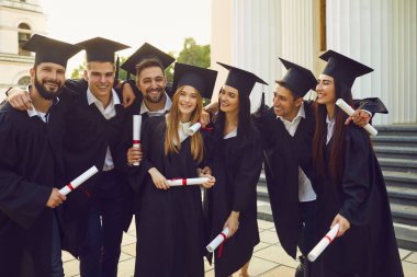 Pozitif öğrencilerden oluşan bir grup üniversite mezunu arkadaşları ayakta duruyor, kucaklıyor ve diplomaları ellerinde tutuyorlar.