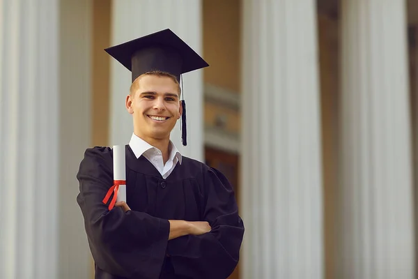 Graduação bem sucedida em vestido acadêmico posando com diploma em mãos contra a formação universitária. — Fotografia de Stock