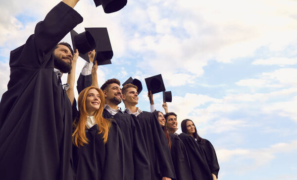Graduates dressed in black mantle standing against a bright sky with raised graduation hats.