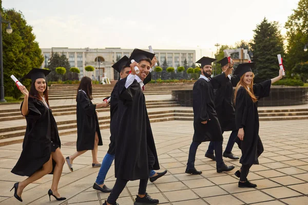 Grupo de jóvenes graduados universitarios sonrientes en mantos tradicionales caminando, con diplomas en manos levantadas — Foto de Stock