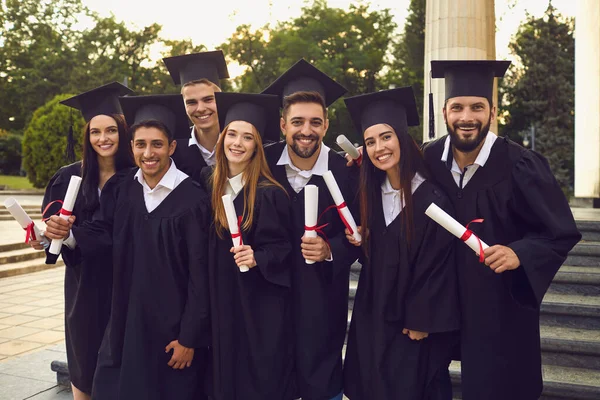 Bir grup mutlu mezun yükseltilmiş ellerle üniversite binasının arka planında yürüyorlar.