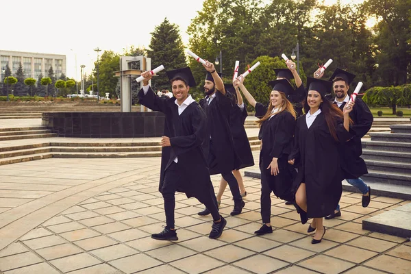 Skupina šťastně se usmívajících absolventů vysokých škol kráčející s diplomy ve zvednutých rukou — Stock fotografie