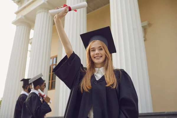 Chica feliz graduada universitaria en manto negro tradicional de pie y con diploma en la mano levantada — Foto de Stock