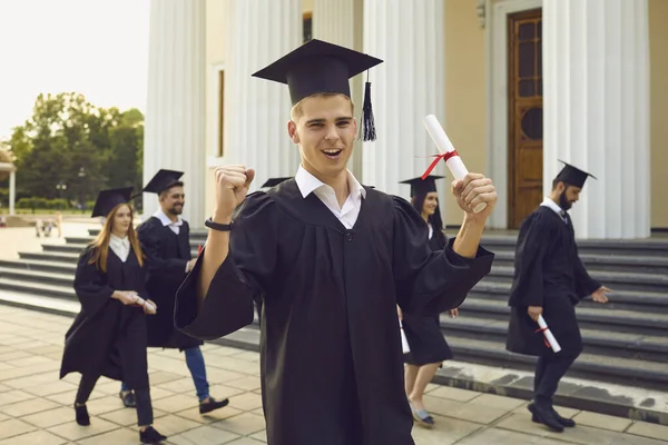 Boldog egyetemi diplomás áll a kezében diploma felett társait diákok séta a háttérben — Stock Fotó