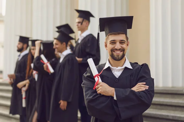 Чоловік у випускних сукнях тримає диплом і посміхається, поки його друзі стоять на задньому плані — стокове фото