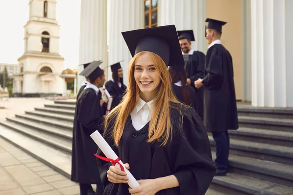 Jong lachend meisje universitair afgestudeerd in traditionele mantel staan en houden diploma in de hand — Stockfoto