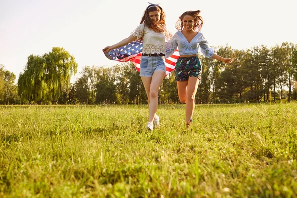 Dívky s americkou vlajkou běhají po trávě v letním parku při západu slunce. 4. července oslava v Americe. — Stock fotografie
