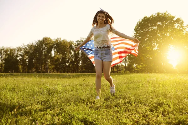 День незалежності США. Дівчинка з американським прапором біжить на траві в літній парку на заході сонця.. — стокове фото