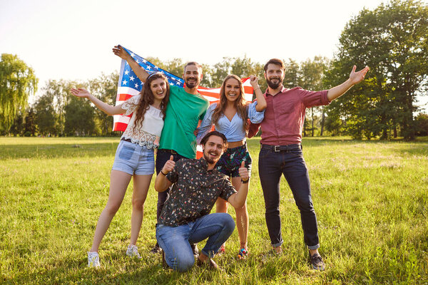 Группа друзей с американским флагом празднуют День независимости Америки, стоя в парке.