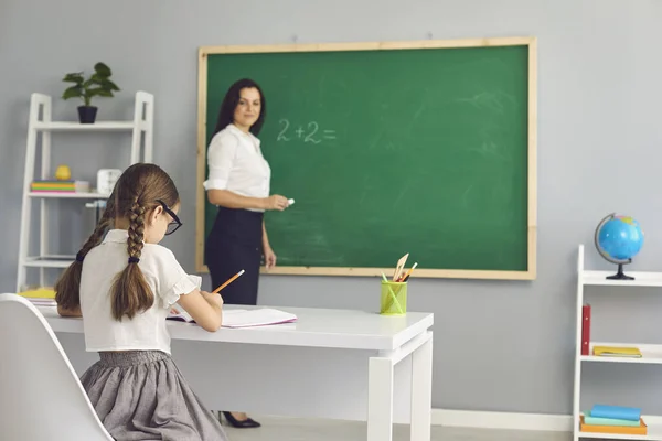 Profesor de escuela primaria en pizarra y colegiala escribiendo sentado en el escritorio — Foto de Stock