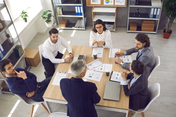 Squadra di giovani lavoratori che ascoltano il senior manager seduto attorno al tavolo dell'ufficio in riunione aziendale — Foto Stock