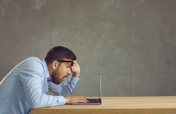 Sorprendido hombre sorprendido con una expresión confusa mira la pantalla del ordenador portátil desde debajo de sus gafas. — Foto de Stock