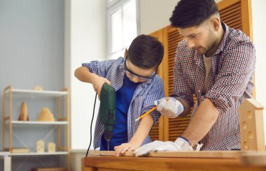 Babasının gözetiminde küçük bir çocuk ev atölyesinde ahşap bir tahta deliyor..