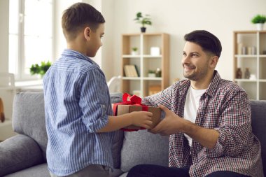 Mutlu gülümseyen baba kanepede oturuyor ve oğullarının ellerinden sürpriz hediyeler alıyor.