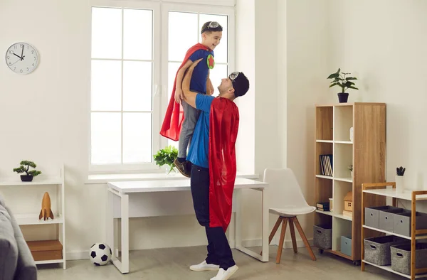 Ευτυχισμένος μπαμπάς και αστείος μικρός γιος ντυμένος με κάπες superhero παίζοντας διασκεδαστικά παιχνίδια στο σπίτι — Φωτογραφία Αρχείου