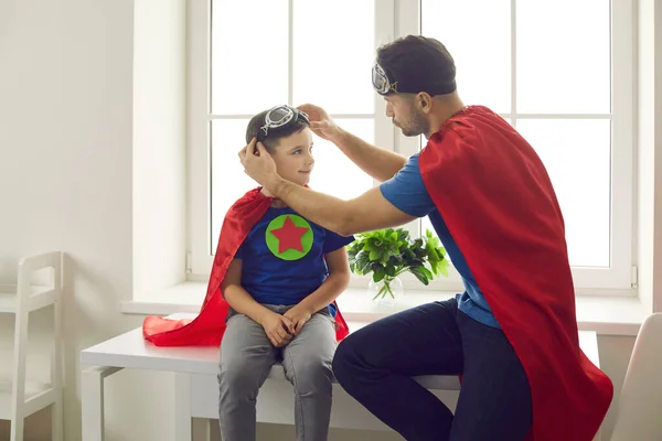 Dad helping son put on aviator goggles while playing fun superhero games at home