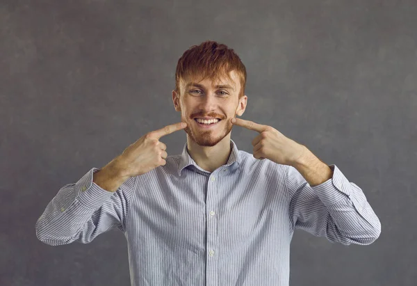 白い歯が灰色の背景に立っているのを示すカジュアルなシャツを着て若い男 — ストック写真