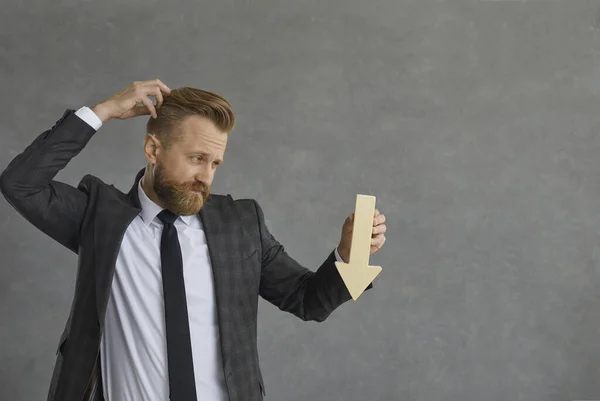 Confused businessman in suit holding arrow down scratching head studio shot — 스톡 사진